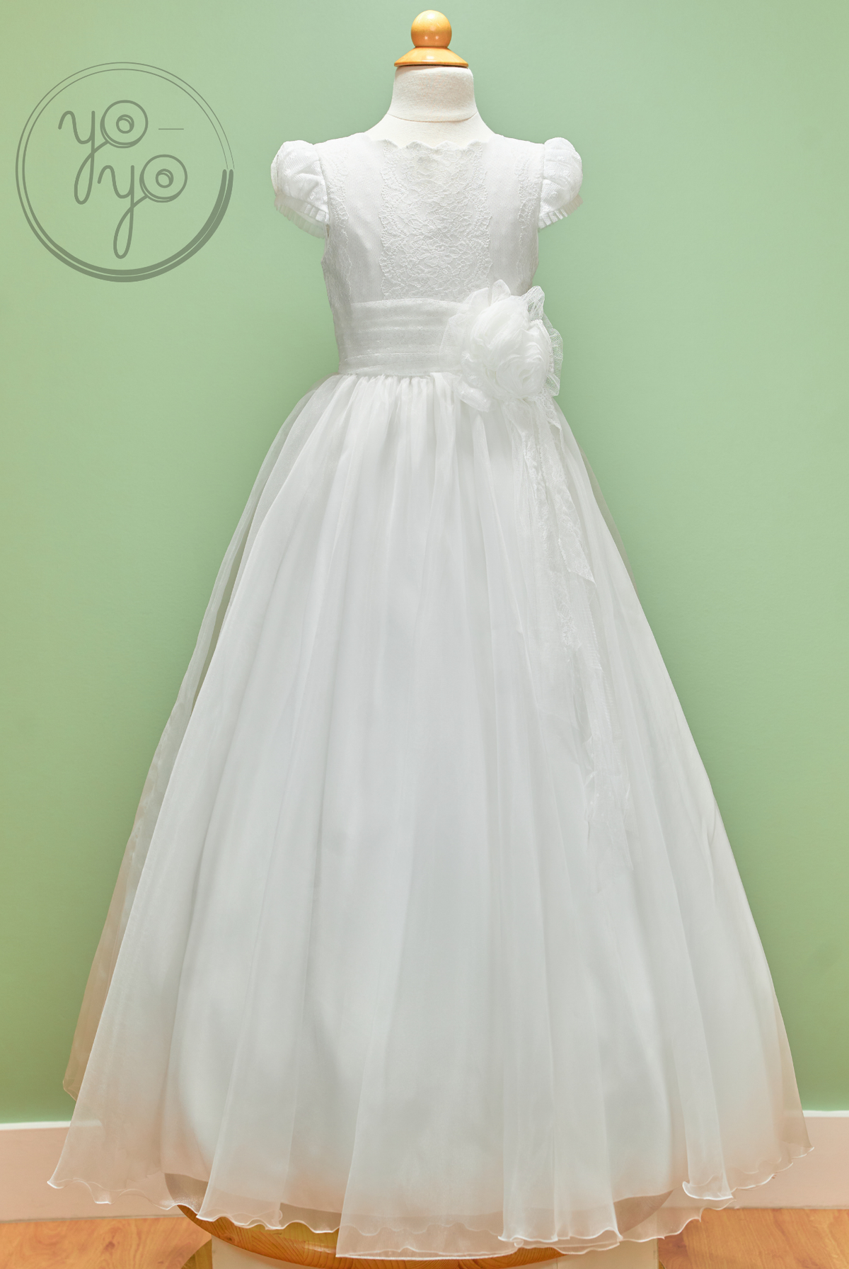 YoYo Boutique Iris Dress on a mannequin: a floor-length white First Communion gown with a lace bodice, puffed sleeves, an organza skirt, and a flower.