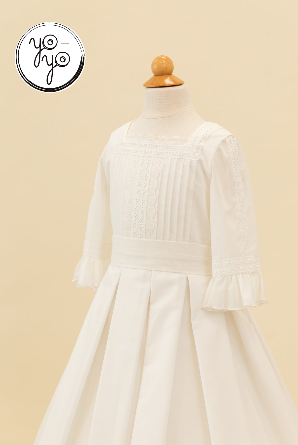White Claudine Dress on a mannequin, featuring a square neckline, lace trim, pintuck bodice, and three-quarter length ruffled sleeves.