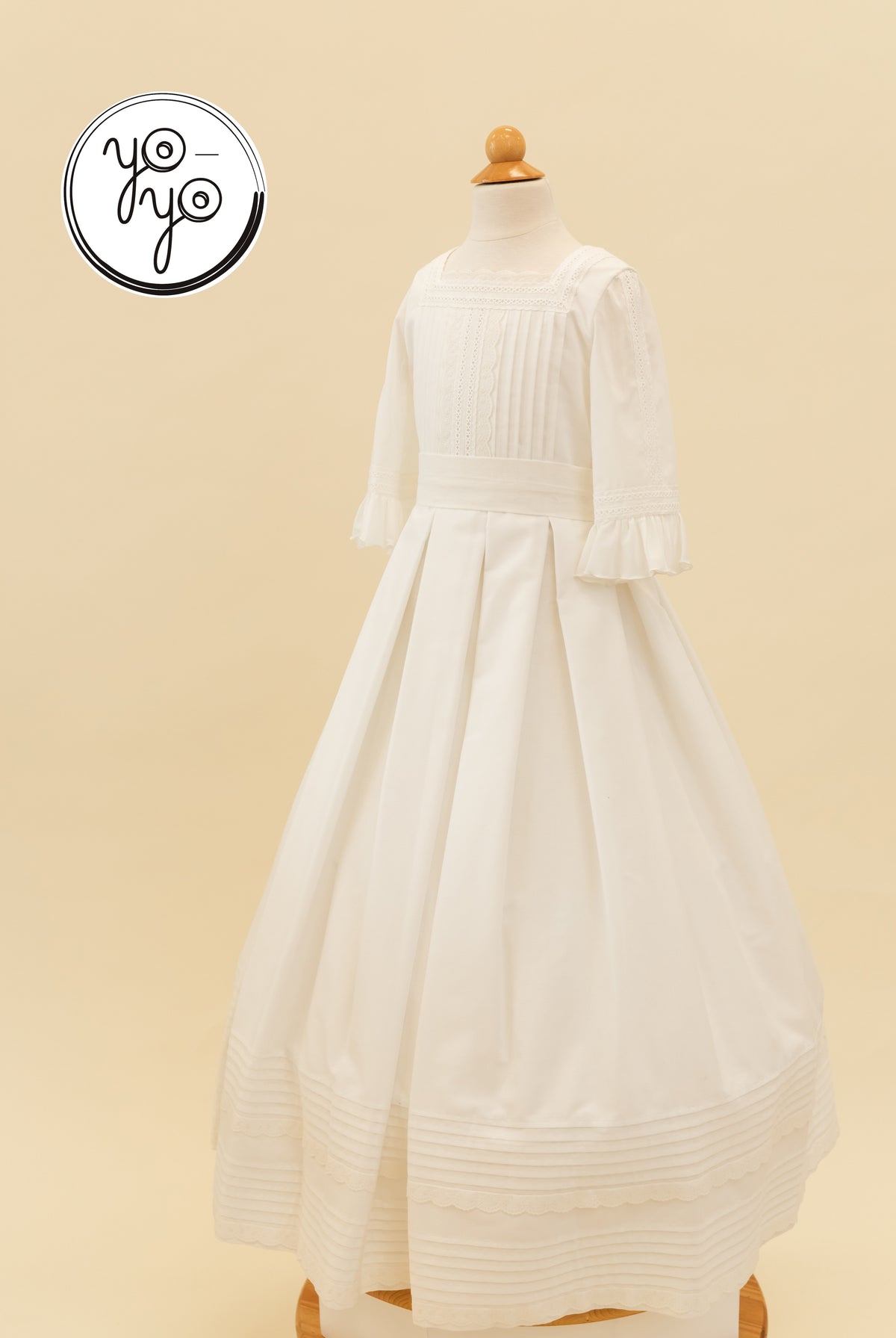 YoYo Boutique’s Claudine Dress: a white floor-length cotton lace gown with a square neckline, pleated bodice, ruffled sleeves, and a tiered hem.