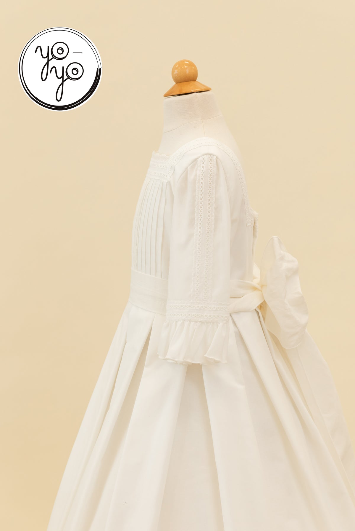 YoYo Boutique Claudine Dress: A white cotton lace gown on a mannequin featuring a pleated bodice, three-quarter sleeves, and a bow.