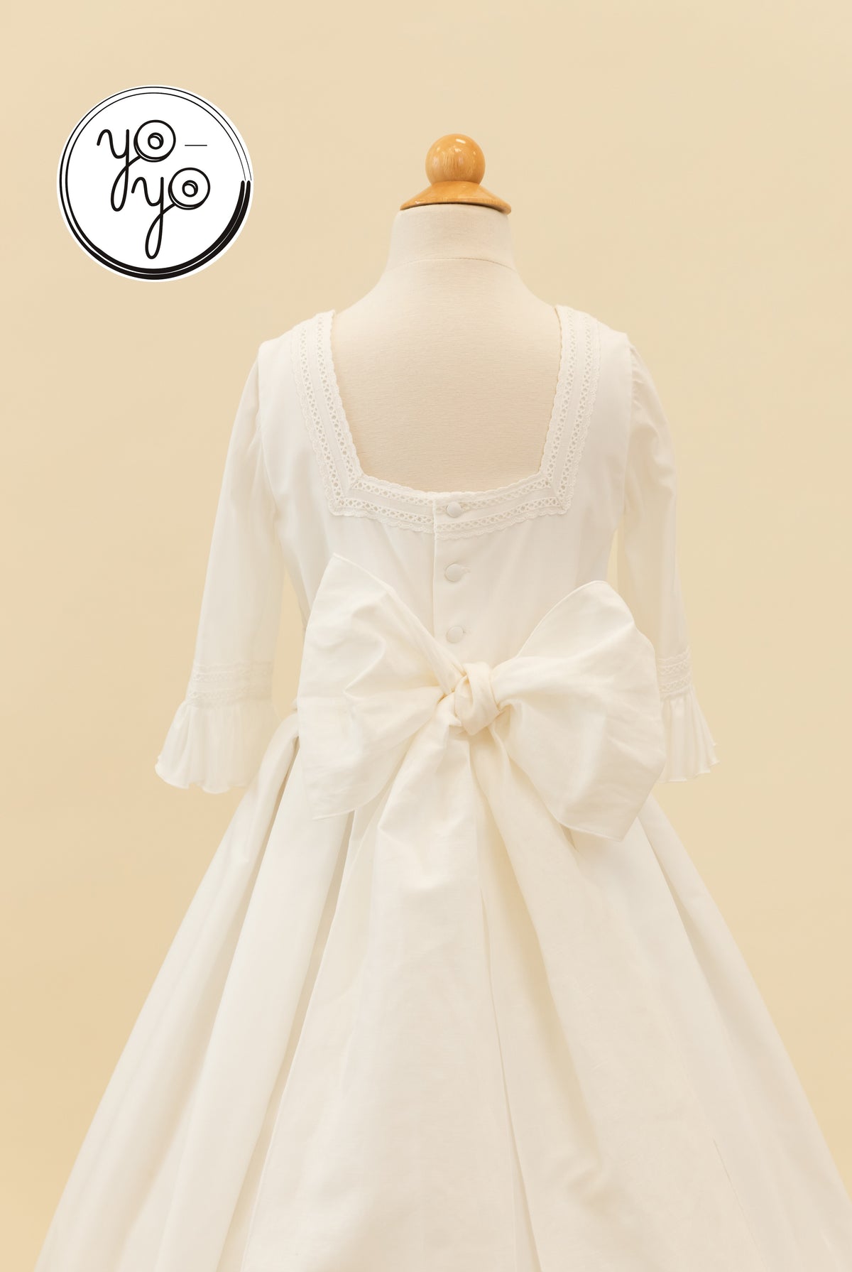 Back view of the Claudine dress in cream cotton lace, featuring a square neckline, ruffled sleeves, a large back bow, and button closures.