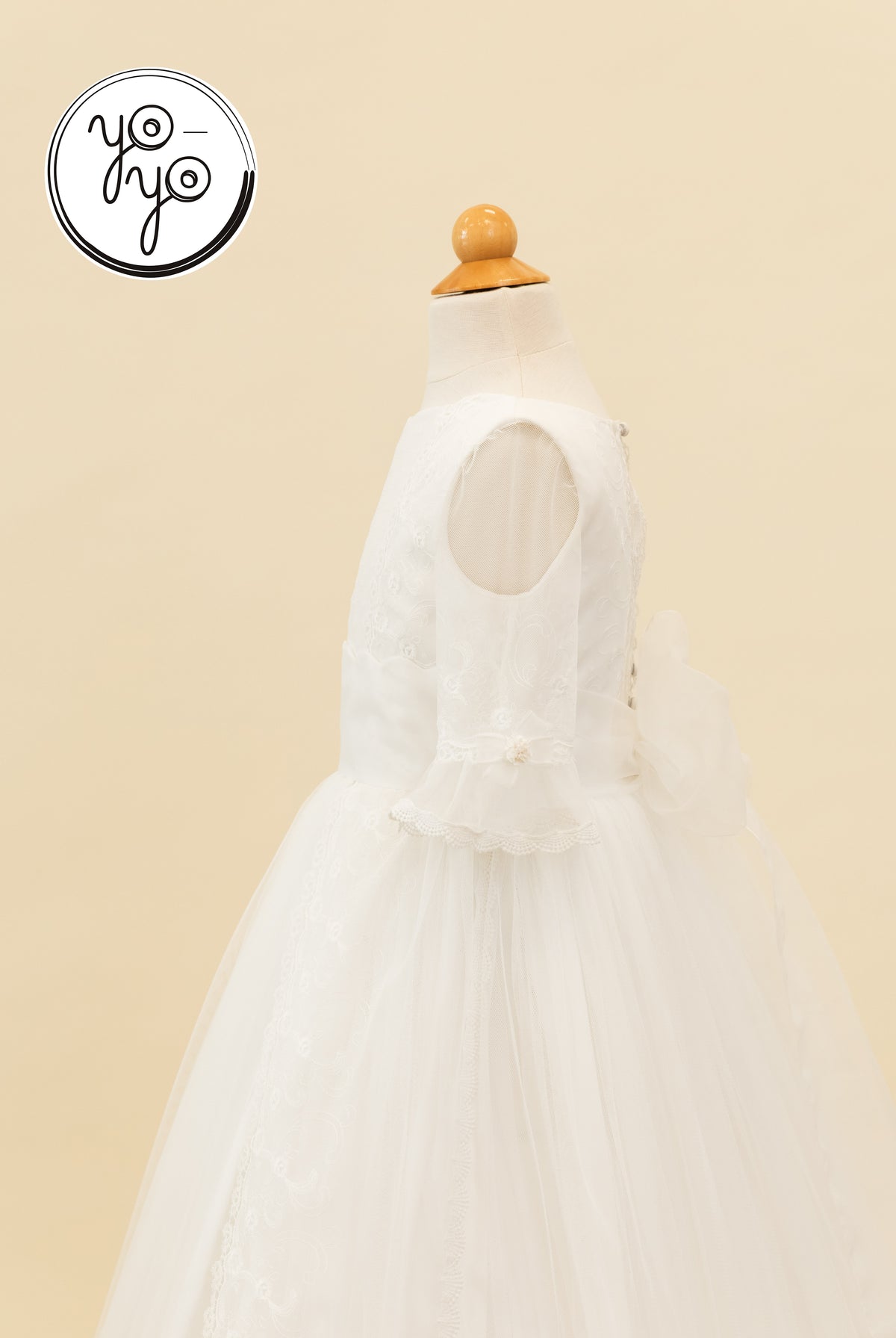 YoYo Boutique Senna white sleeveless boho Communion dress on a mannequin, featuring lace overlay, flutter sleeves, and a large back bow.