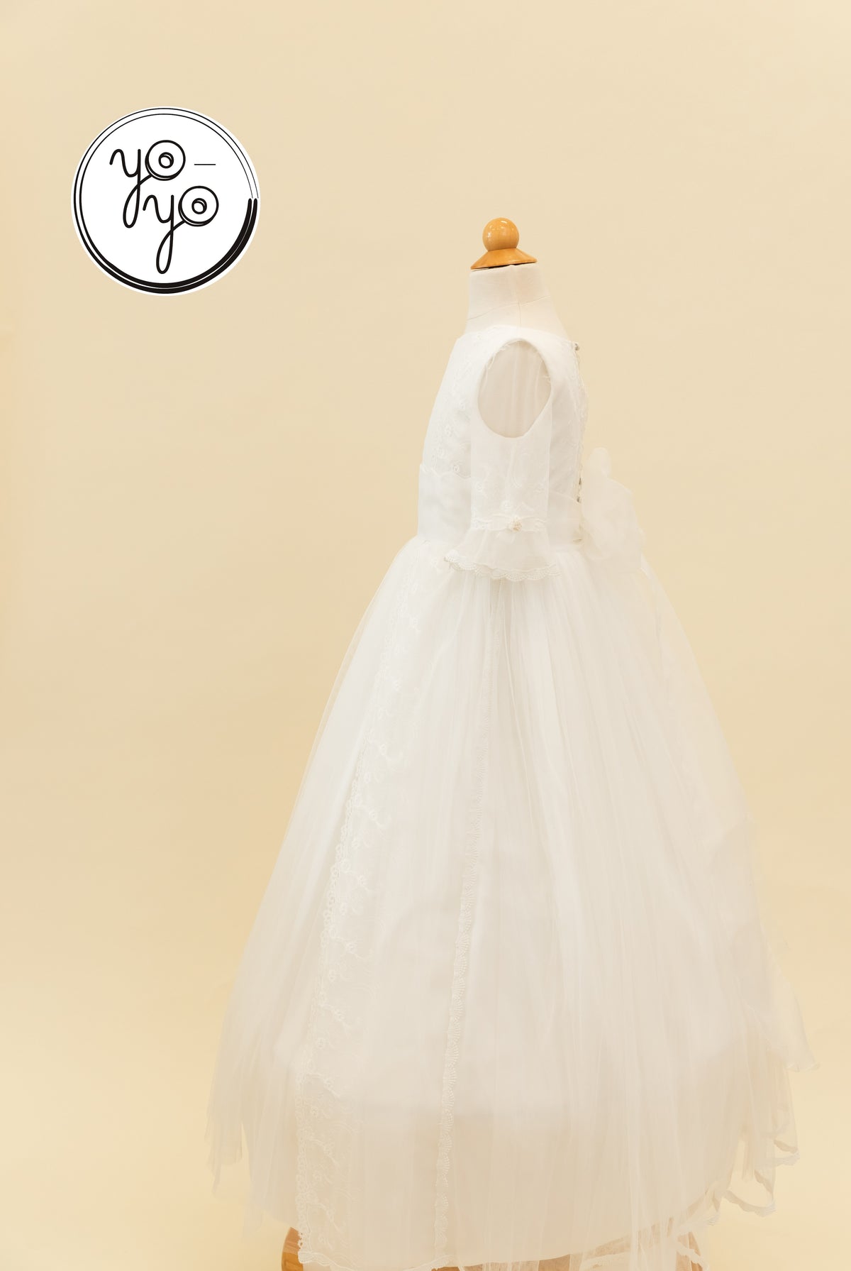 YoYo Boutique Senna Dress: a white, floor-length First Communion gown featuring a sheer overlay, ruffled sleeves, and delicate lace detailing.