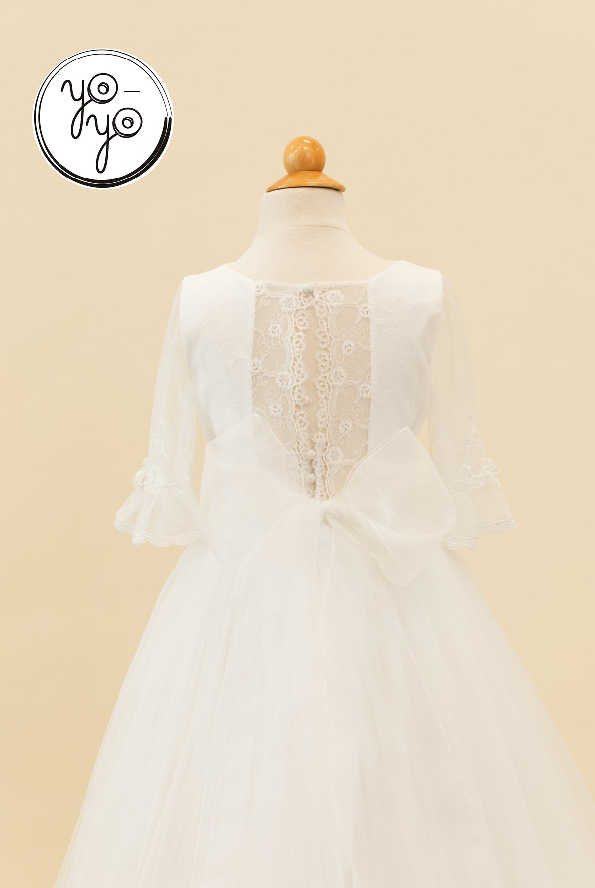 Back view of the white boho Senna Dress featuring sheer lace sleeves, a lace back panel with button closures, and a large organza waist bow.
