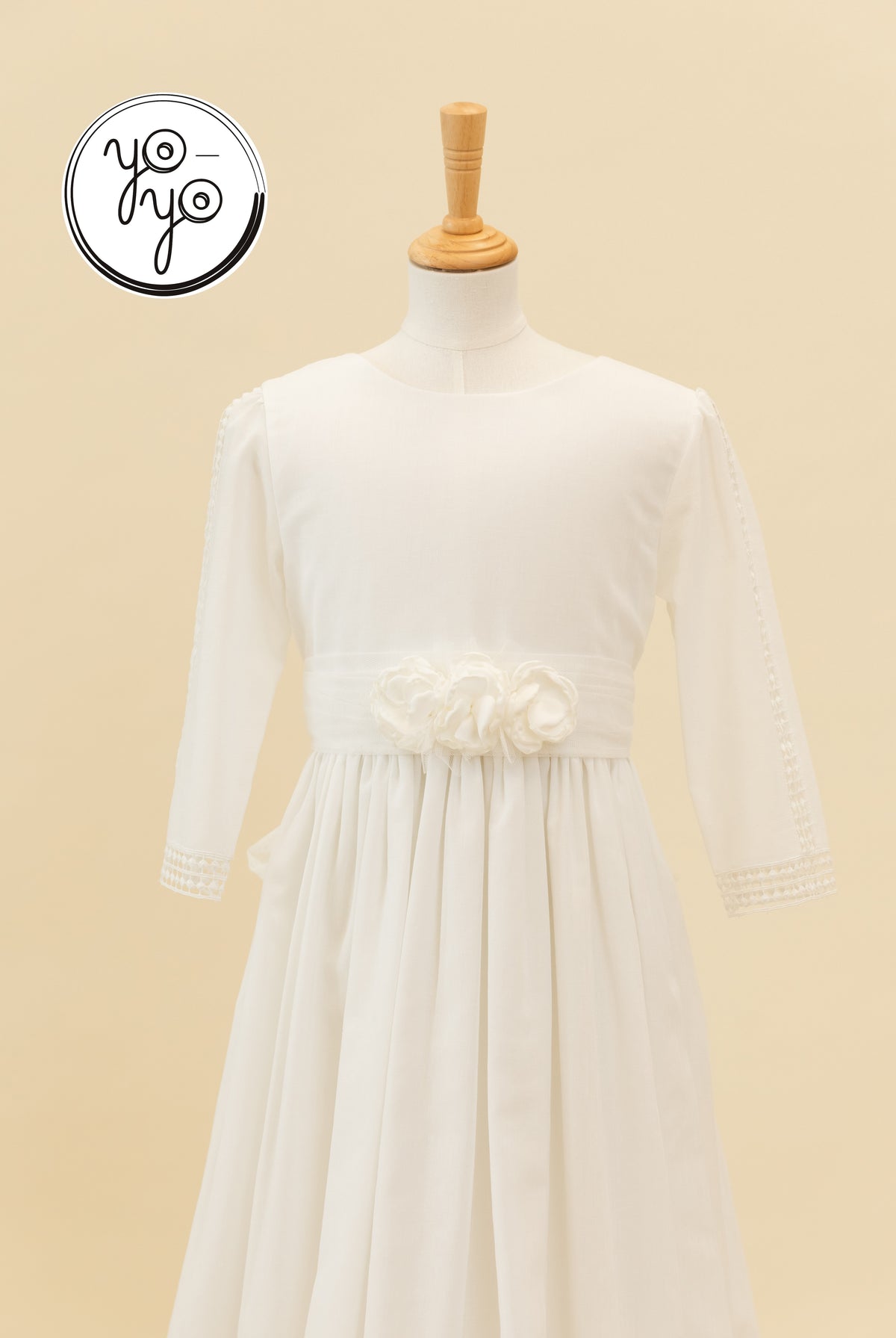 White linen Montserrat dress featuring a round neck, wide waistband with three fabric flowers, and lace trim on the sleeves and hem.