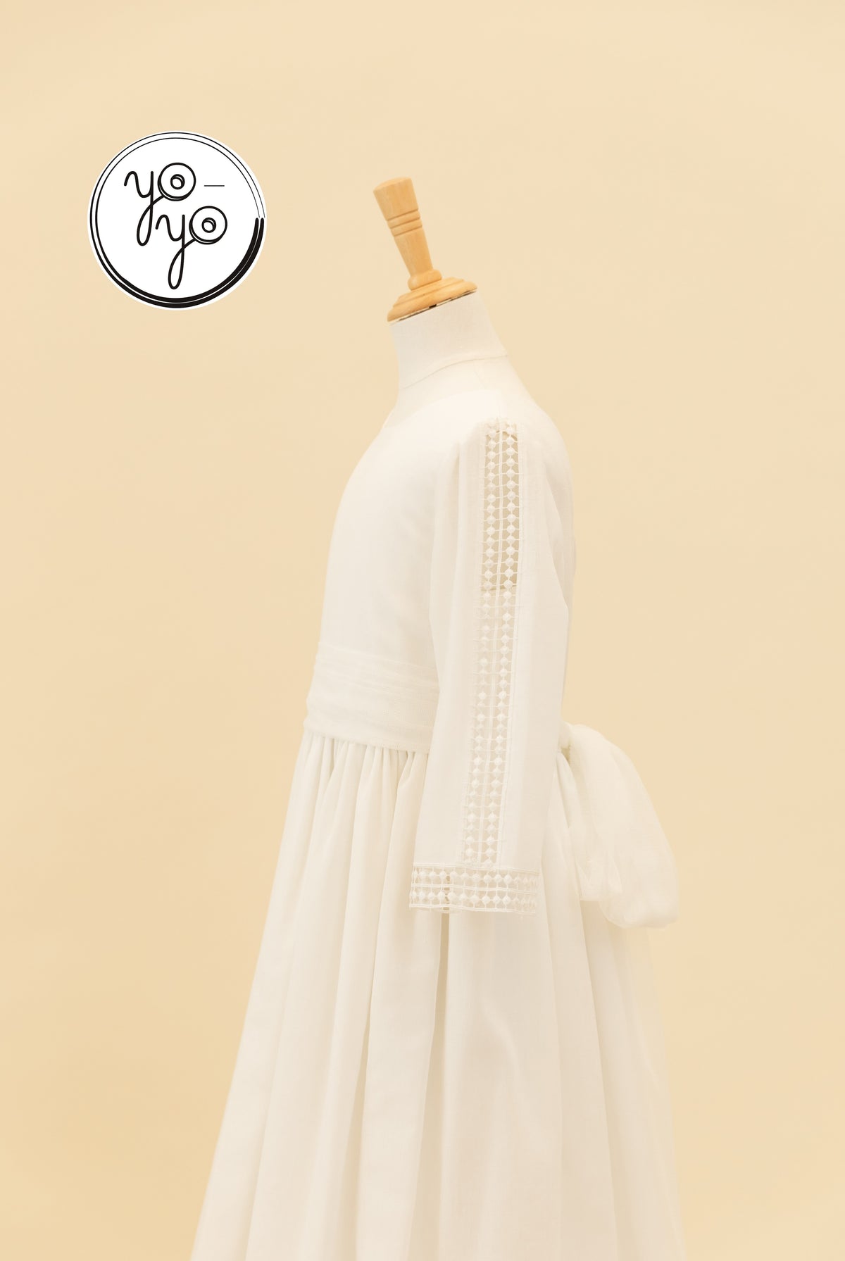 YoYo Boutique Montserrat cream linen communion dress on a mannequin, featuring long lace sleeves and a tie-back waist.