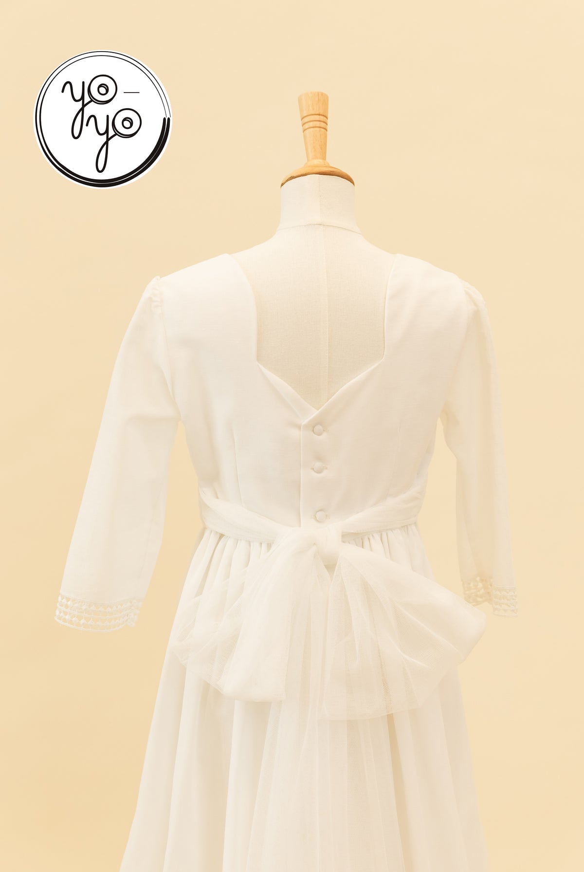 Back view of the white linen Montserrat Dress featuring a V-back, button closure, lace-trimmed three-quarter sleeves, and a large tulle waist bow.
