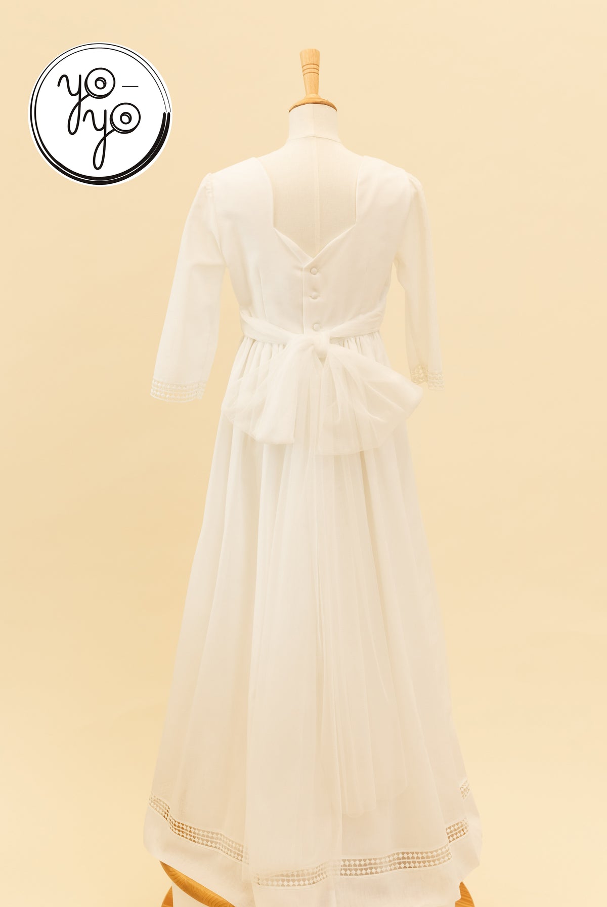 Back view of the white linen Montserrat Dress, featuring three-quarter sleeves with lace trim, a large tulle waist bow, and a lace-trimmed hem.