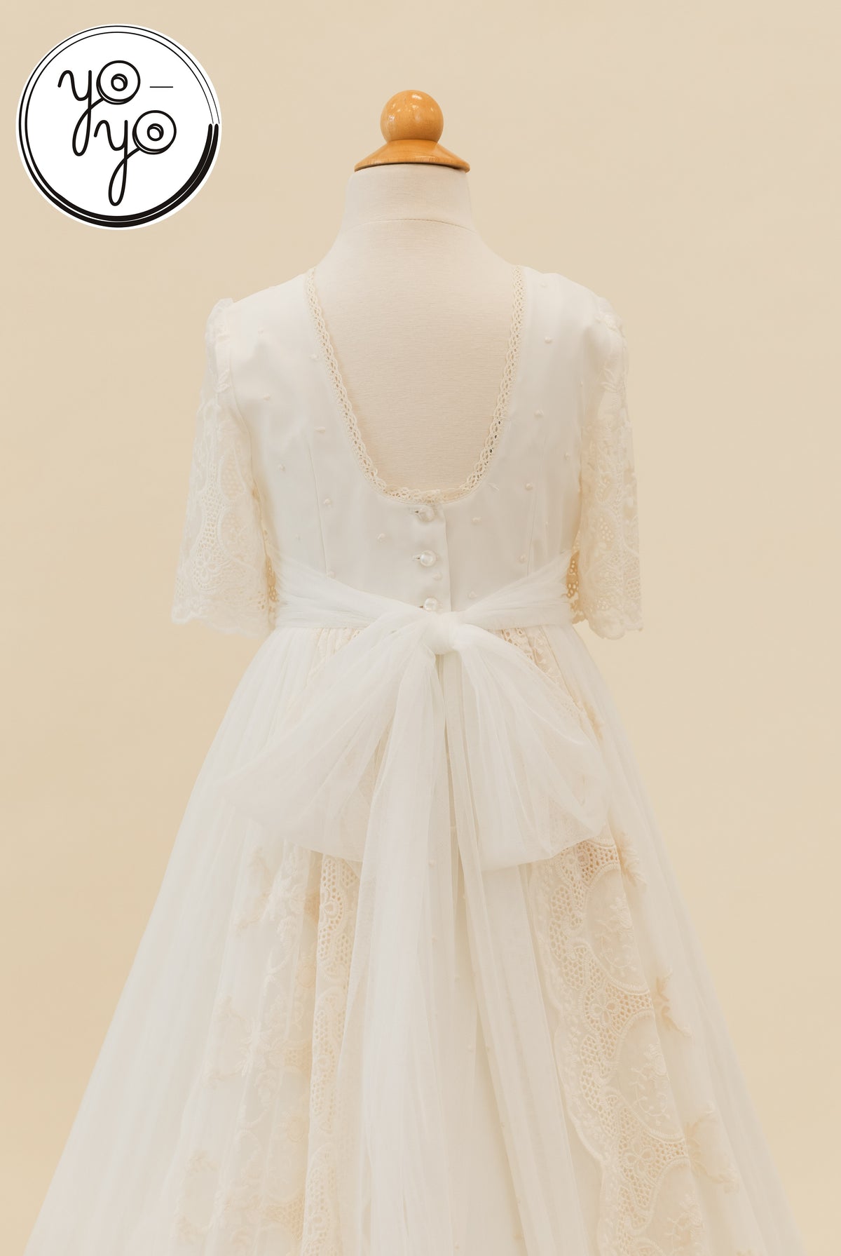 Back view of the white boho Pino Dress by YoYo Boutique, featuring a lace-trimmed scoop neckline, button back, and a large tulle bow.