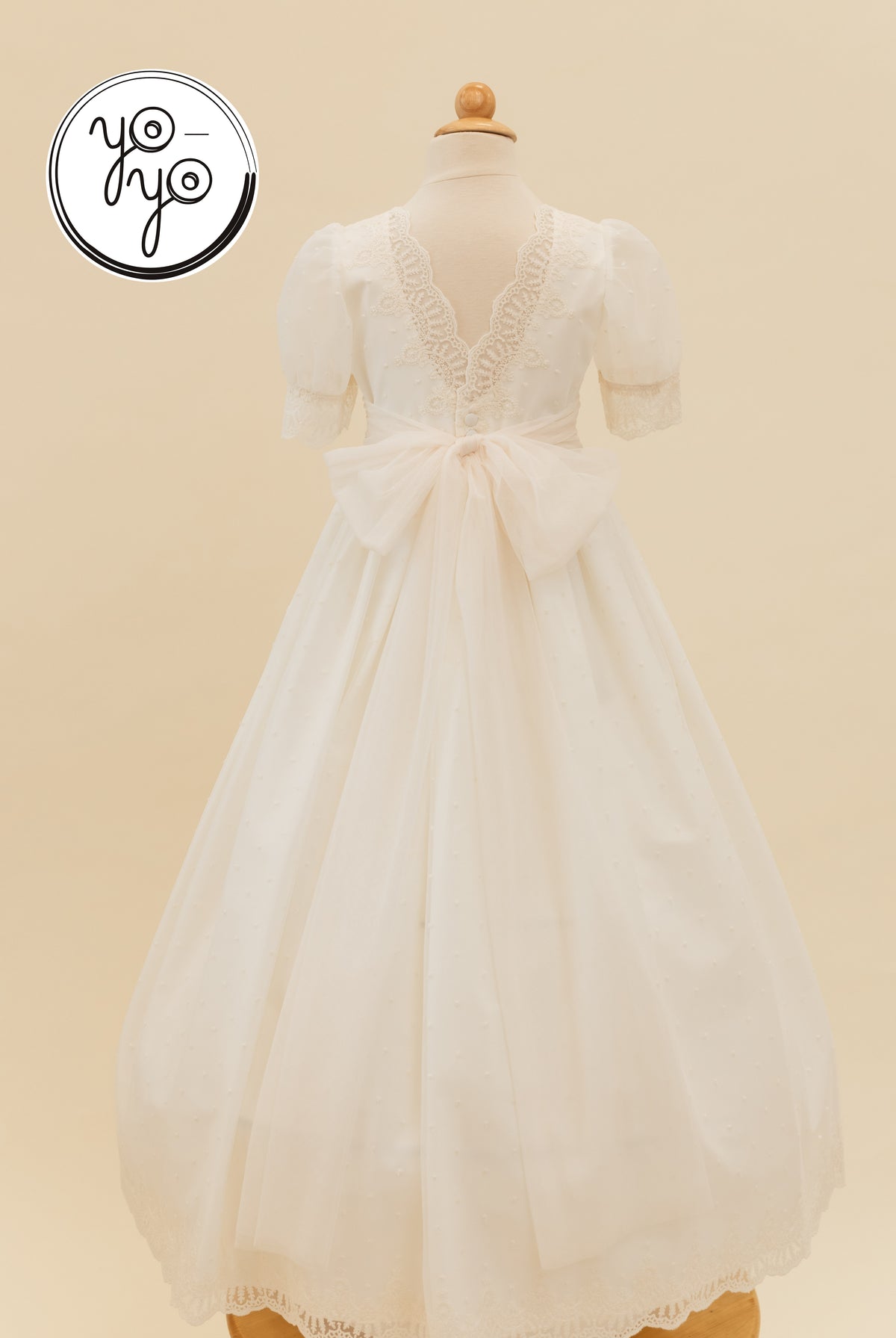 Back view of the white, floor-length Sophie Dress by YoYo Boutique, featuring puff sleeves, lace trim, and a large sheer back bow.