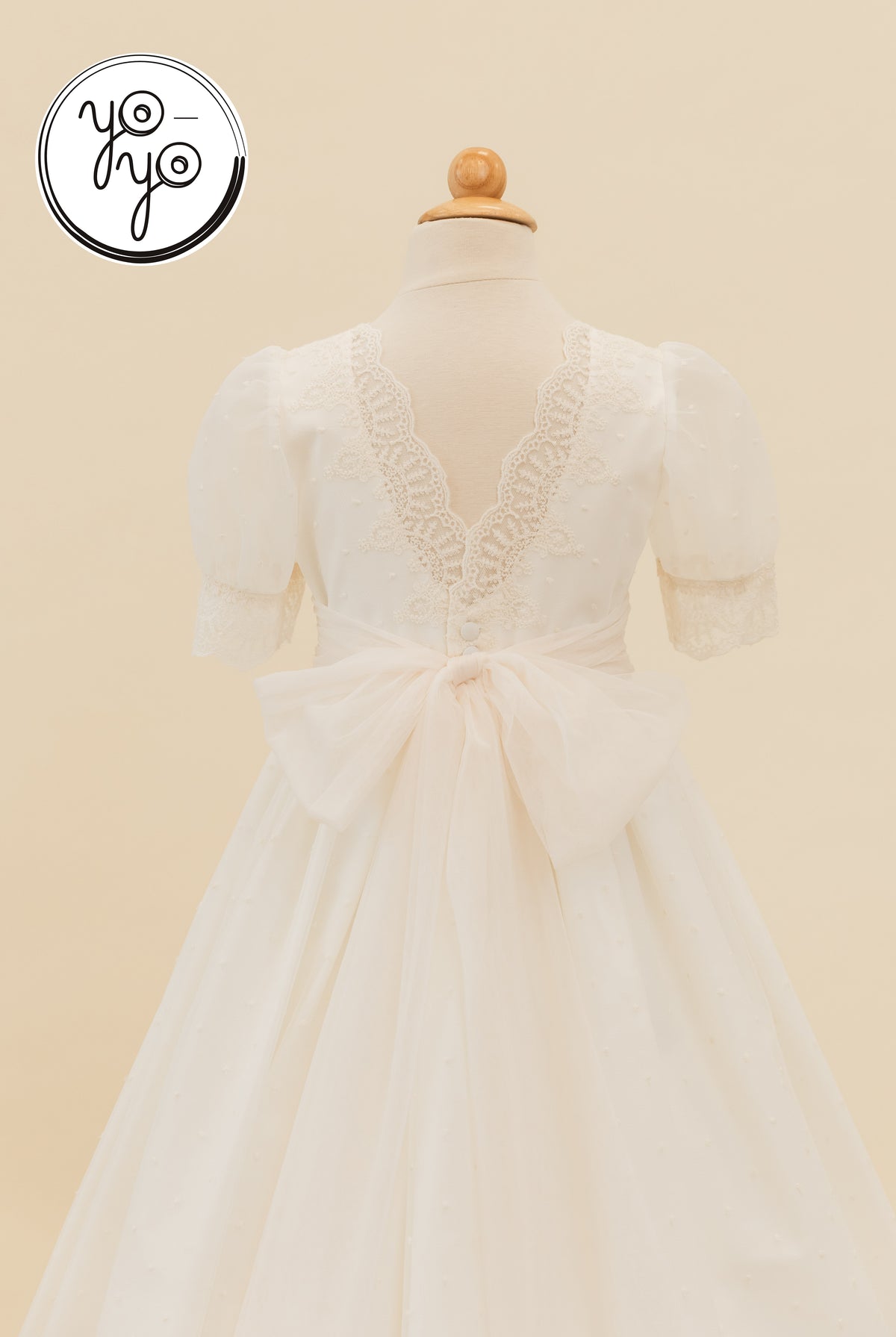 Back view of the white, short-sleeved YoYo Boutique Sophie Dress with a V-shaped lace back, button detail, and large sheer waist bow.