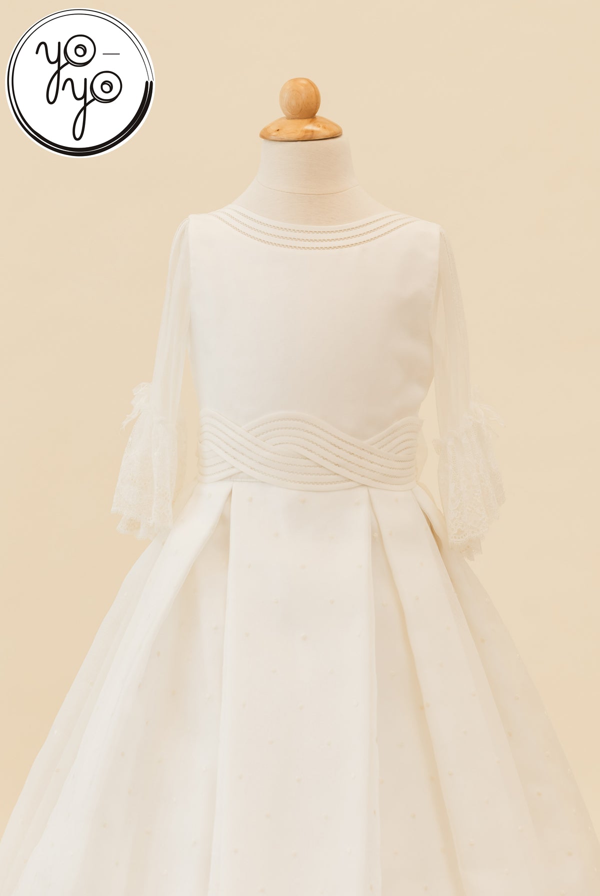 Off-white Ceibo A-line First Communion gown on a mannequin featuring a boat neckline, sheer lace sleeves, and a wavy-stitched waist.