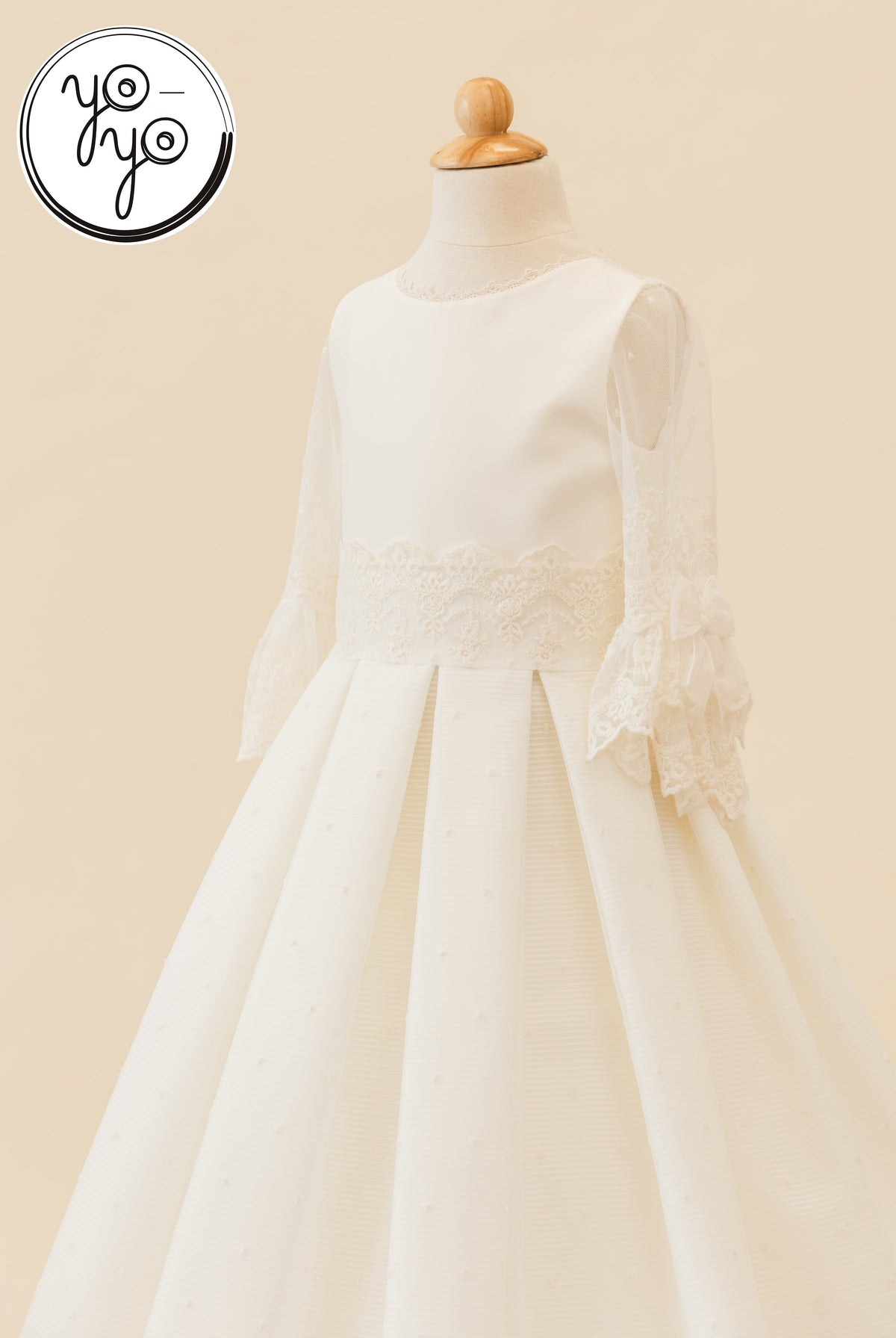 Cream YoYo Boutique Giselle Deluxe dress on a mannequin, featuring long sleeves, lace, a pleated skirt, and an embroidered tulle pattern.