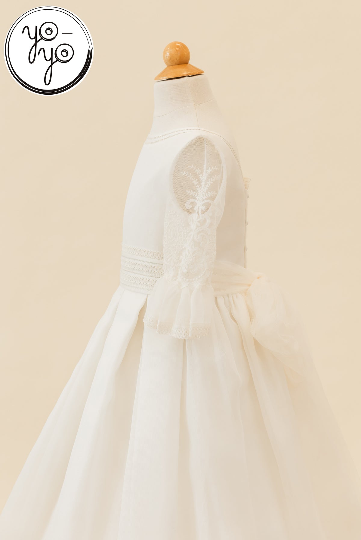 Side view of a white Magnolia Dress on a mannequin, featuring embroidered lace sleeves and a large back waist bow.