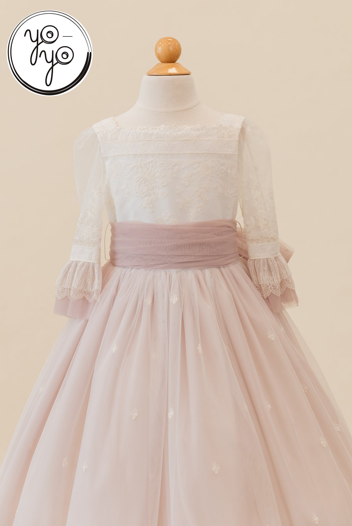 The Elena Dress by YoYo Boutique is a boho Communion gown with a white embroidered bodice, sheer sleeves, and a dusty pink tulle skirt with a sash.