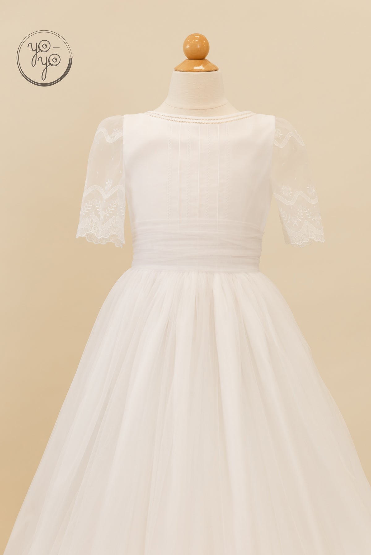 The Lunaria Dress by YoYo Boutique is a white First Communion dress featuring lace short sleeves, a sheer tulle overlay, and a draped waist.