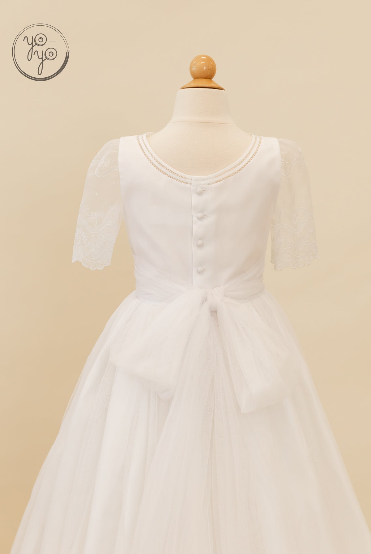 Back view of the white Lunaria Dress by YoYo Boutique, featuring sheer lace sleeves, a button row, and a large tulle bow at the waist.