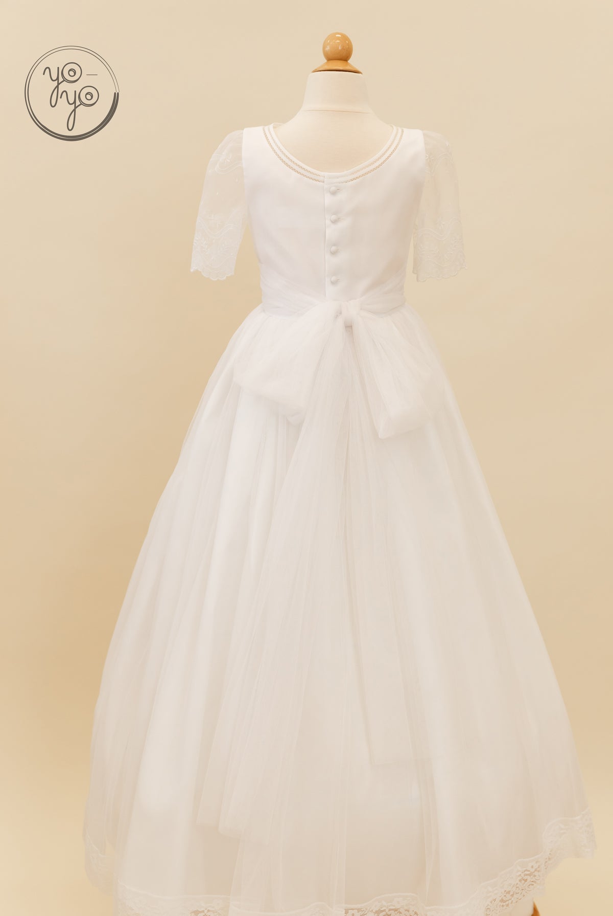 Back view of the white organza Lunaria Dress by YoYo Boutique with sheer lace sleeves, a button-up back, and a tulle skirt with a waist bow.
