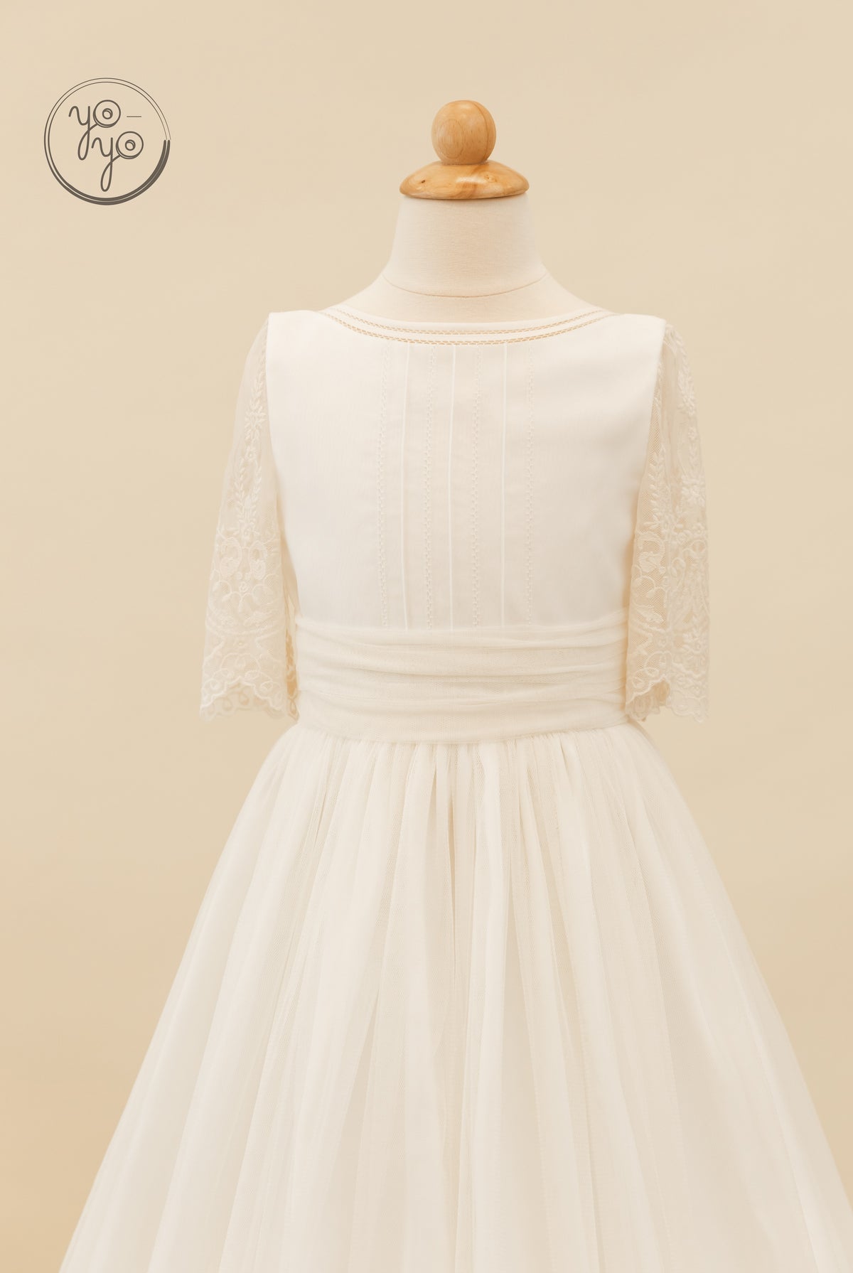 Cream Melisa Dress on a mannequin, featuring a pleated bodice, elbow-length lace sleeves, and a layered tulle skirt.