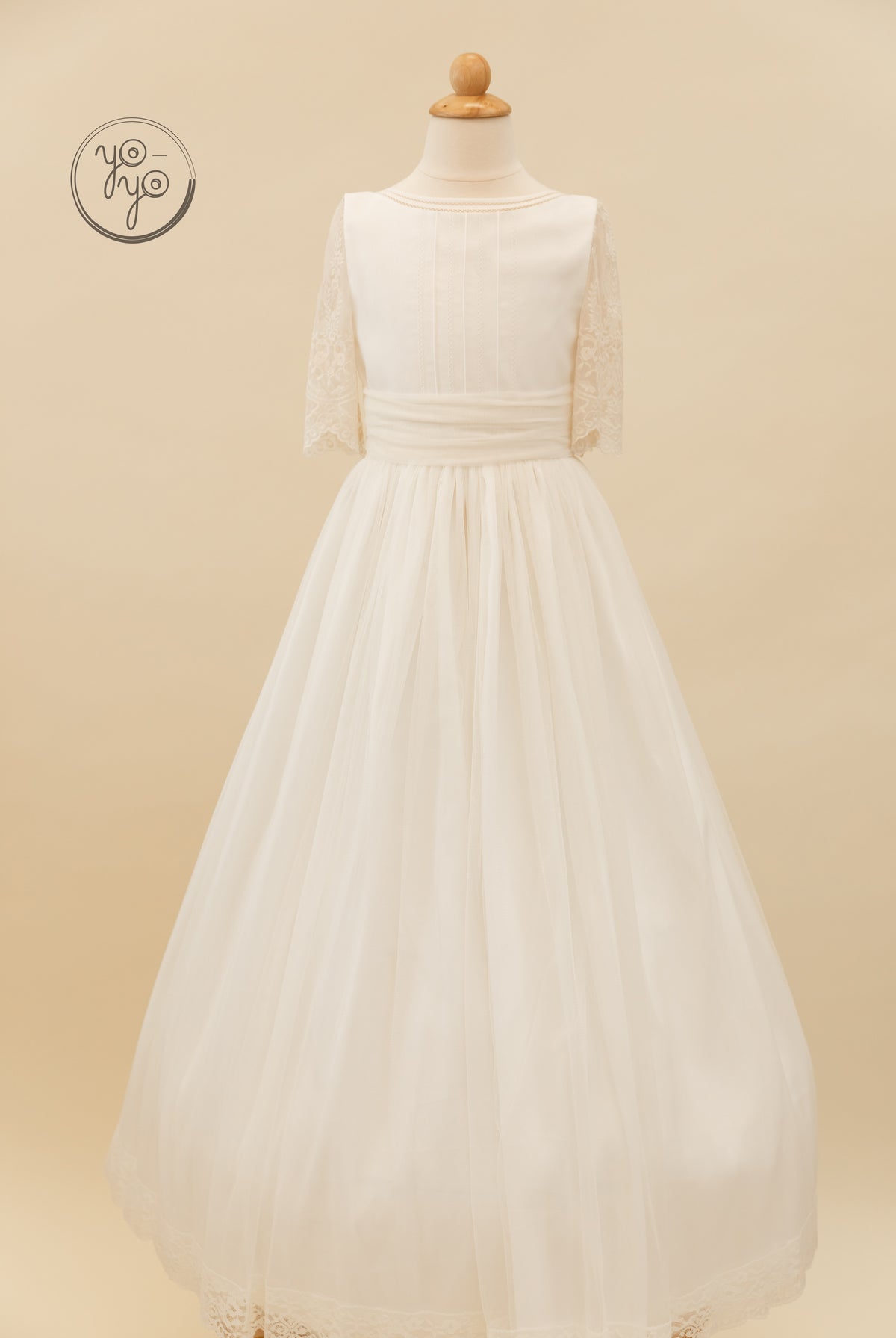 Cream Melisa Dress with a pleated bodice, elbow-length lace sleeves, and a floor-length tulle skirt with a lace-trimmed hem.