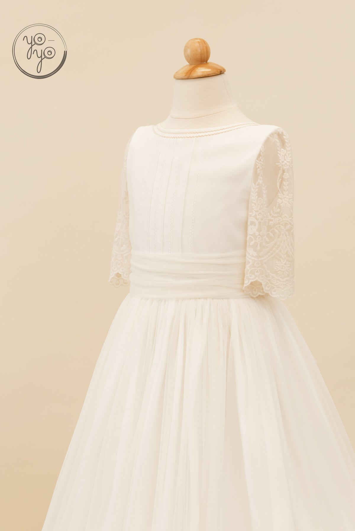 Melisa Dress by YoYo Boutique: off-white First Communion dress on a mannequin featuring a pleated bodice, wide sash, lace sleeves, and tulle skirt.