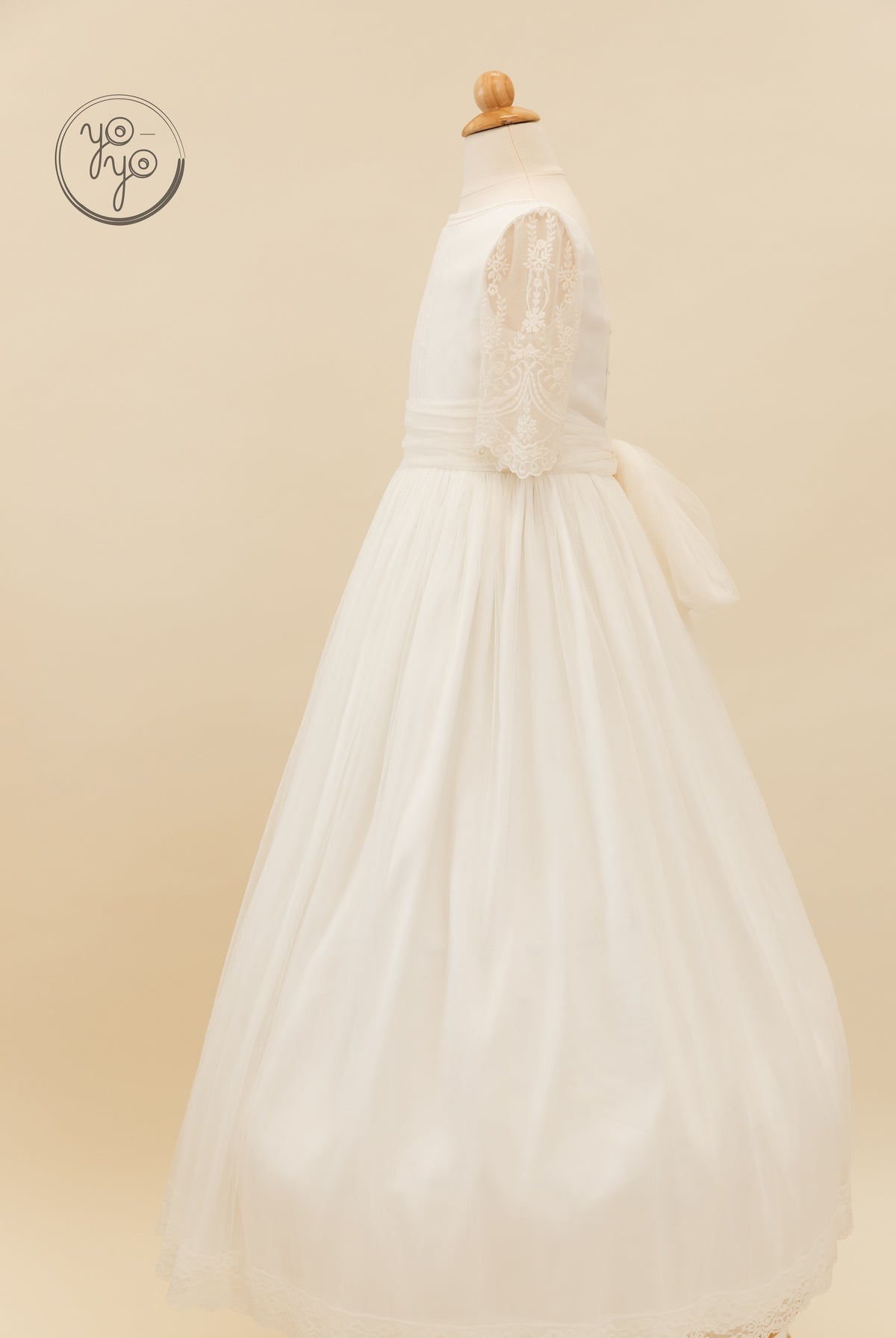 The Melisa Dress by YoYo Boutique: a white, floor-length boho communion dress on a mannequin featuring lace sleeves, a pleated sash, and tiered skirt.