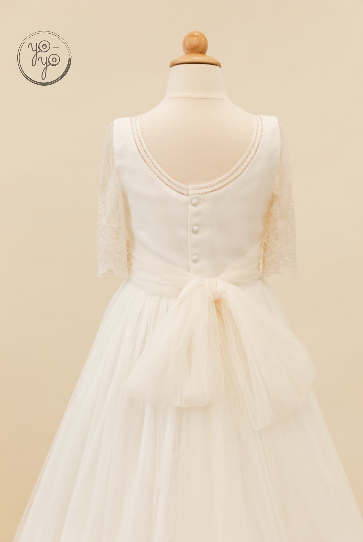 Back view of the white lace and tulle Melisa Dress, featuring a scoop back, button closure, lace sleeves, and a large tulle waist bow.