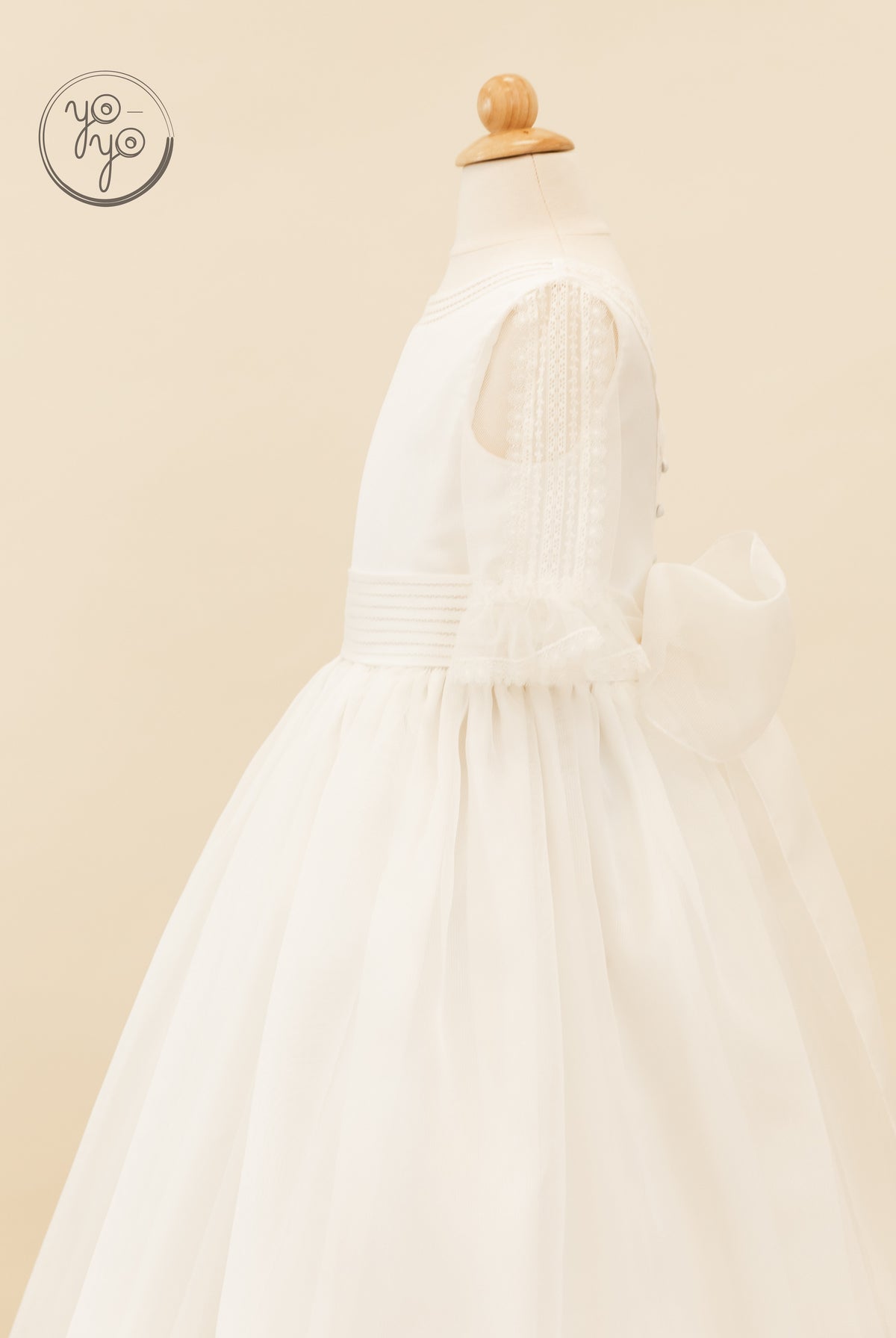 Side view of a white boho Jacinto dress featuring sheer lace sleeves, a wide pleated waistband, and a large back bow.