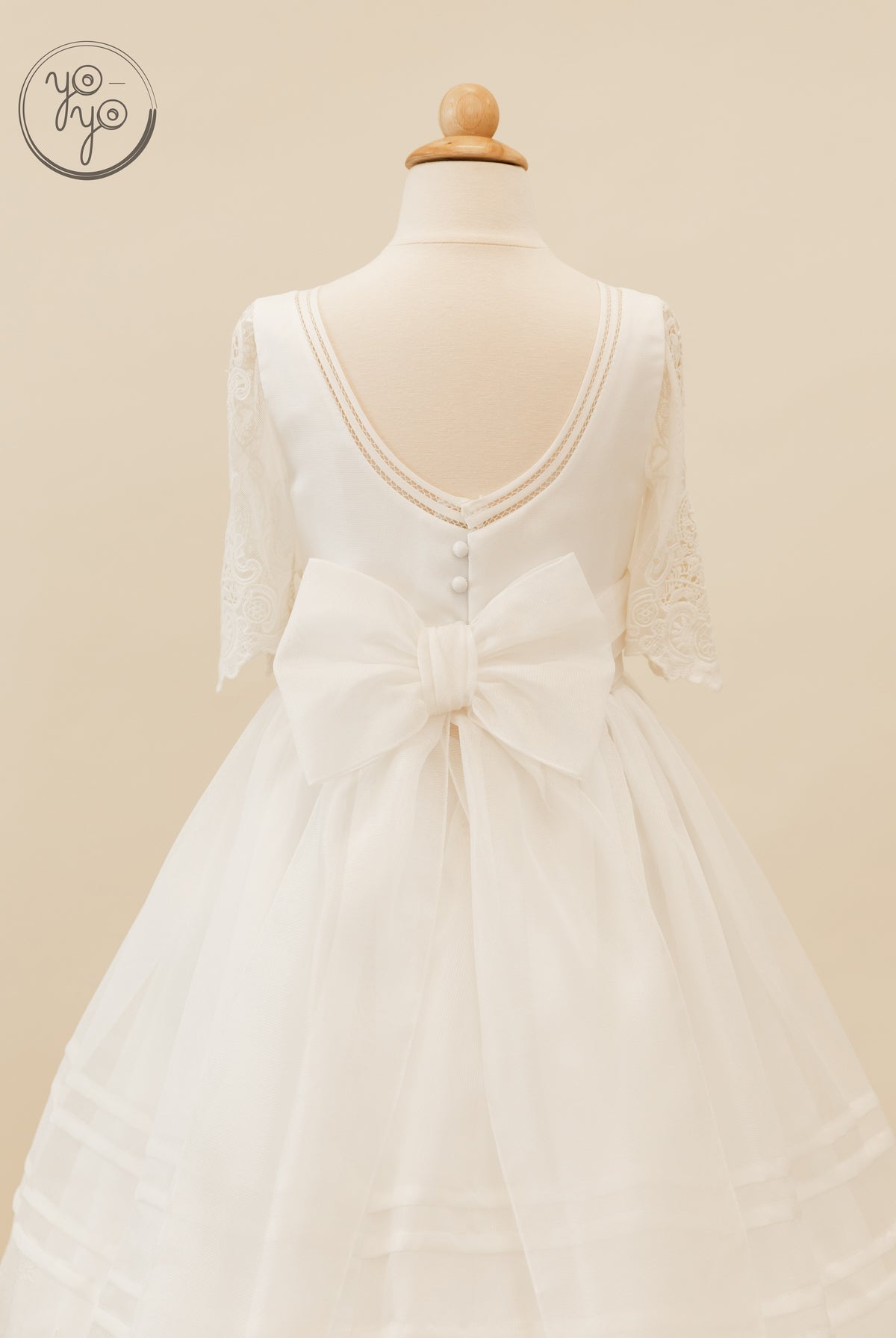 Back view of the white embroidered Coral Dress featuring a scoop neck, lace elbow-length sleeves, button details, and a large waist bow.