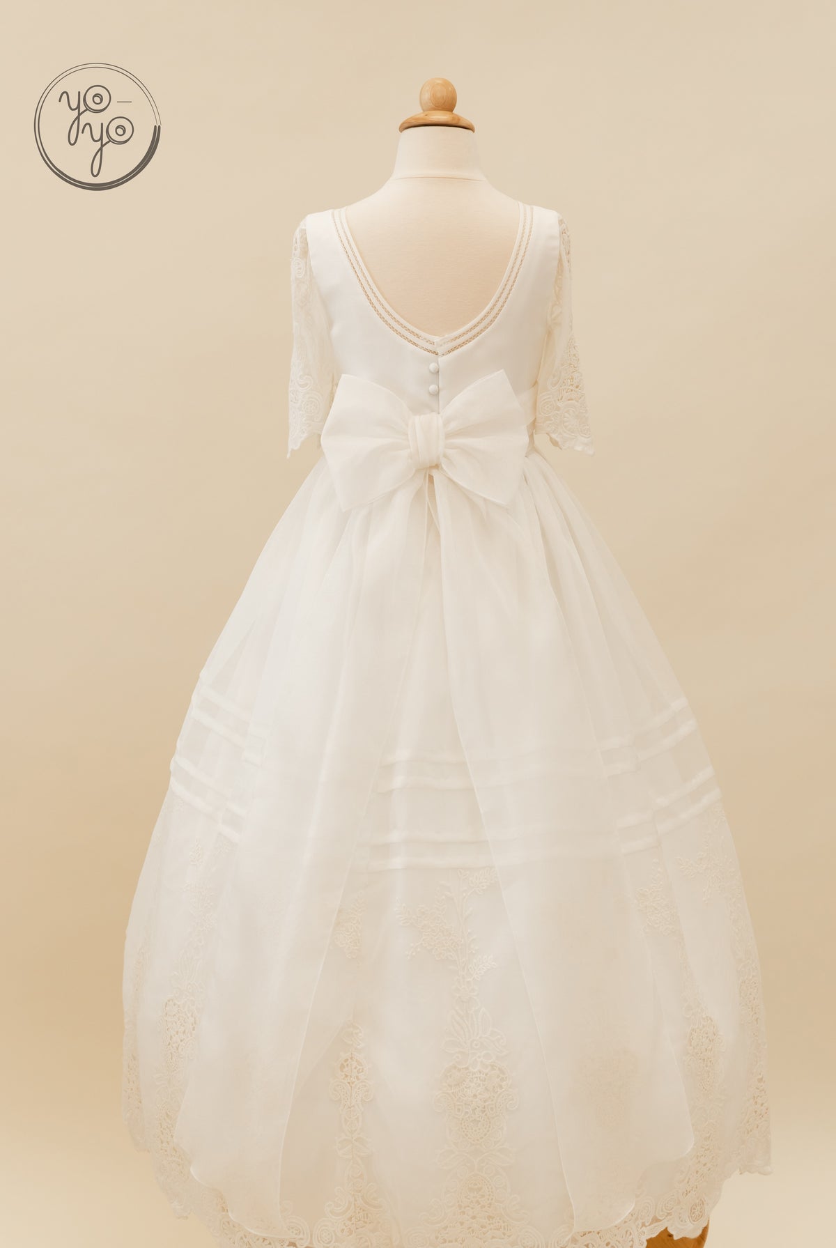 Back view of a white embroidered Coral Dress on a mannequin, featuring lace sleeves, a V-neckline, a large back bow, and a lace-trimmed hem.
