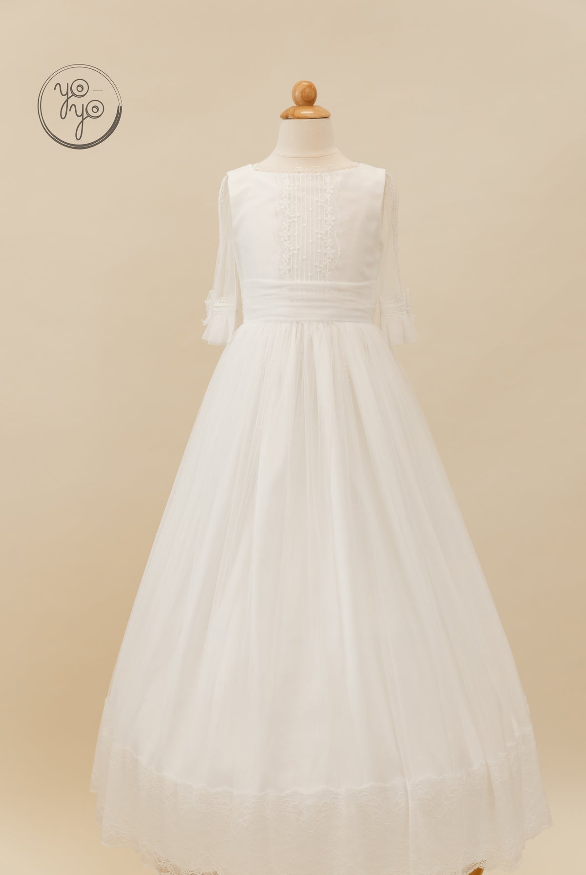 White floor-length Valeriana dress on a mannequin, featuring a pleated bodice, sheer three-quarter sleeves, and a lace-trimmed hem.