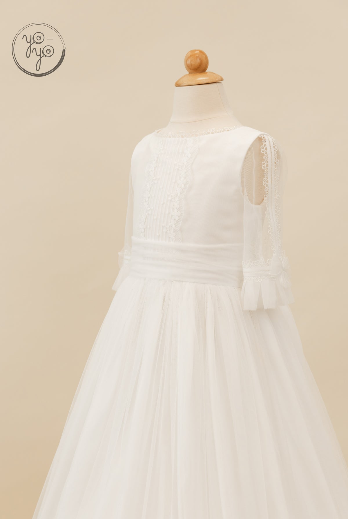 Valeriana Dress by YoYo Boutique: a white French tulle First Communion dress on a mannequin with sheer sleeves, lace trim, and pleated bodice panels.