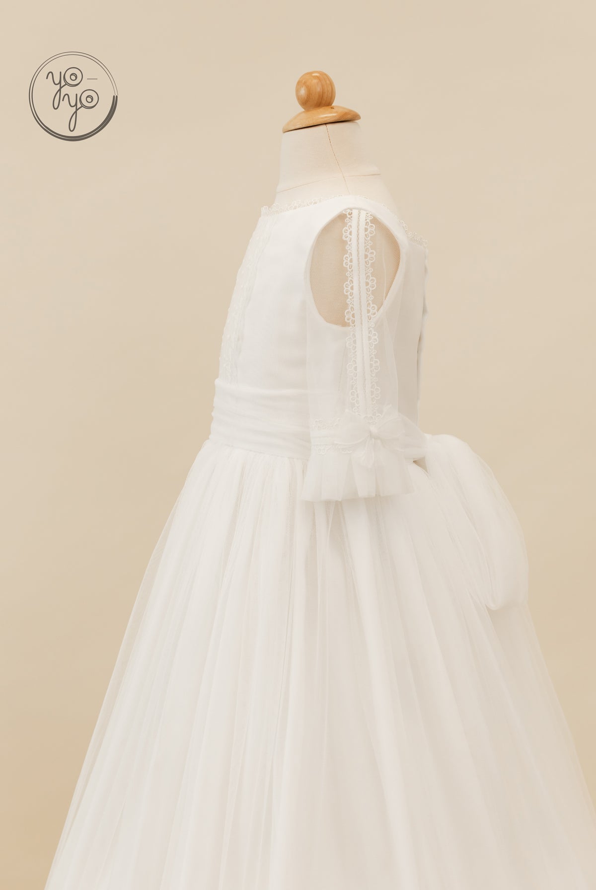 White Valeriana Dress by YoYo Boutique on a mannequin, featuring a pleated bodice, lace-trimmed sheer sleeves, and a large back waist bow.