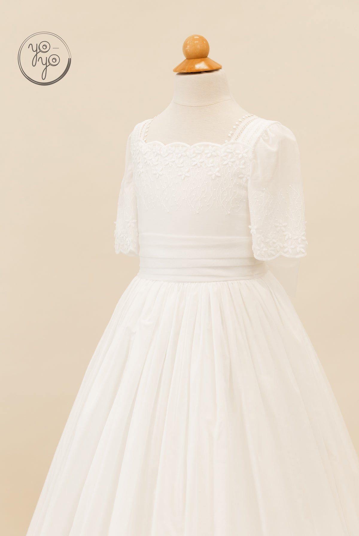 YoYo Boutique Salvia white First Communion dress on a mannequin featuring a scalloped floral embroidered neckline, wide waistband, and full skirt.