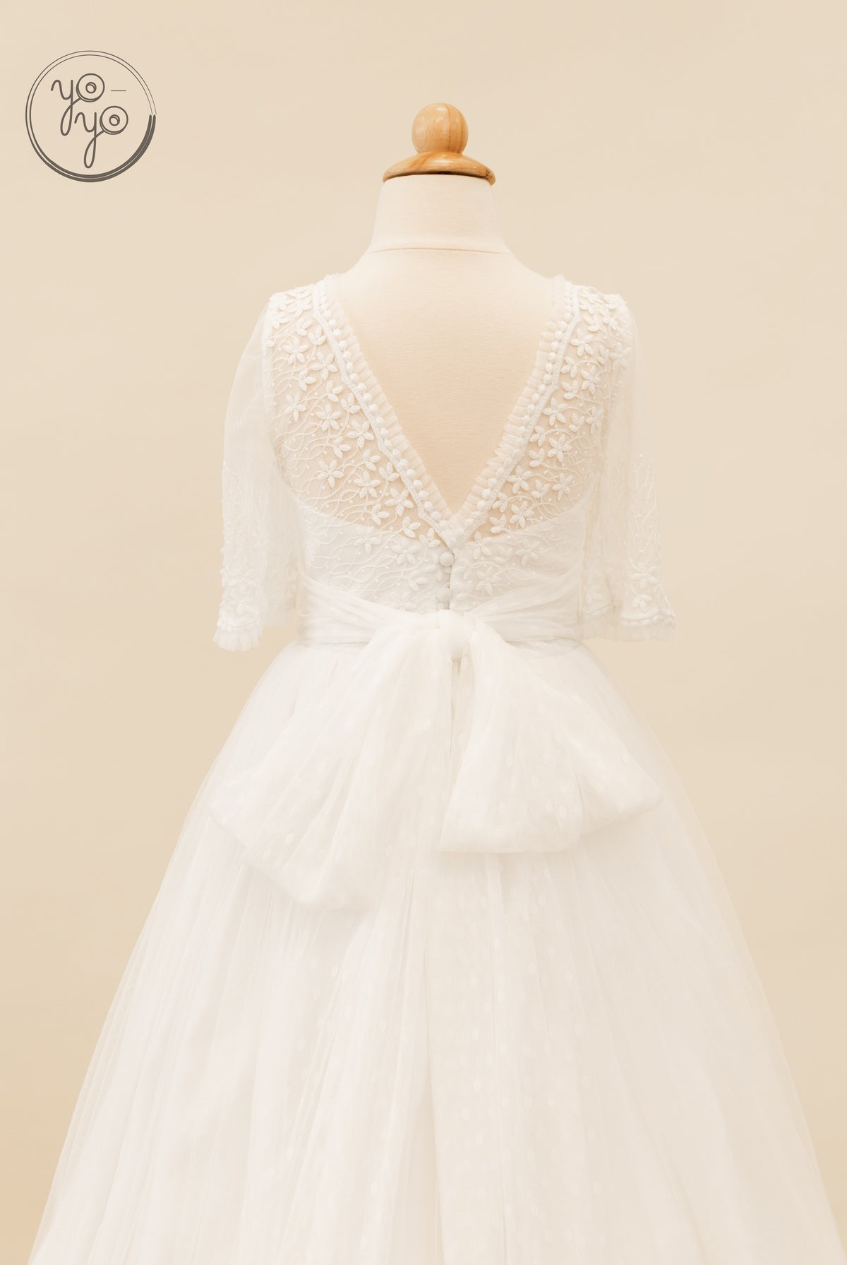 Back view of the white Hibisco Dress with floral embroidery, sheer lace sleeves, a V-back, button details, and a large waist bow.