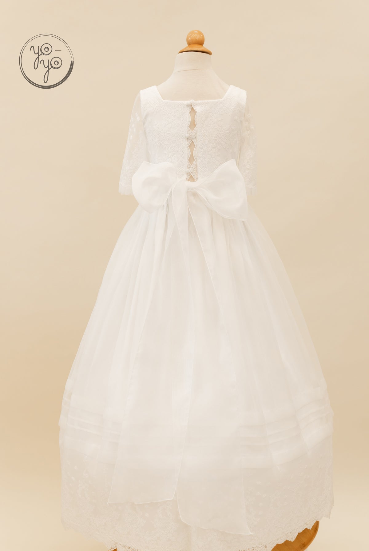 Back view of a white Gladiola Dress with lace sleeves, a square neckline, and a large bow at the waist over a full, pleated skirt.
