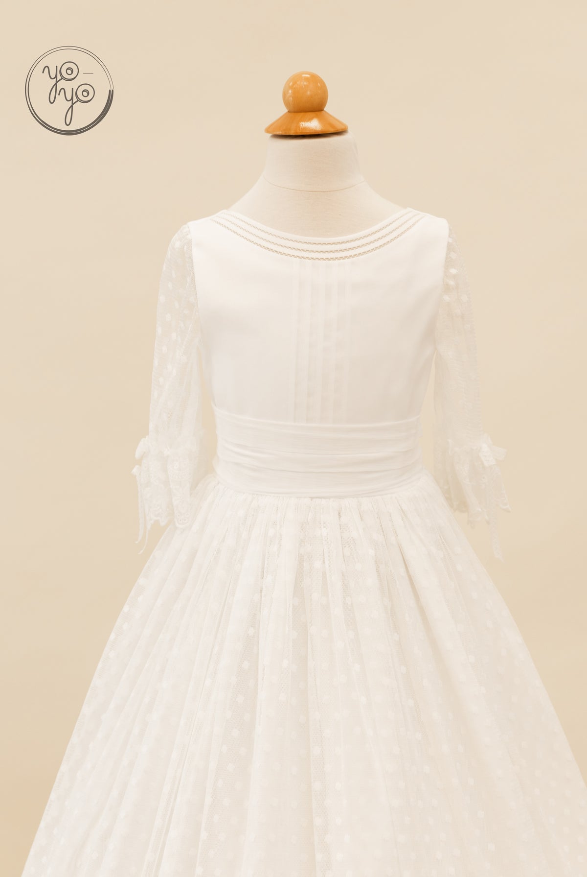 White boho Cassia First Communion dress featuring a wide waistband, sheer three-quarter sleeves with bow cuffs, and a pleated, trimmed bodice.