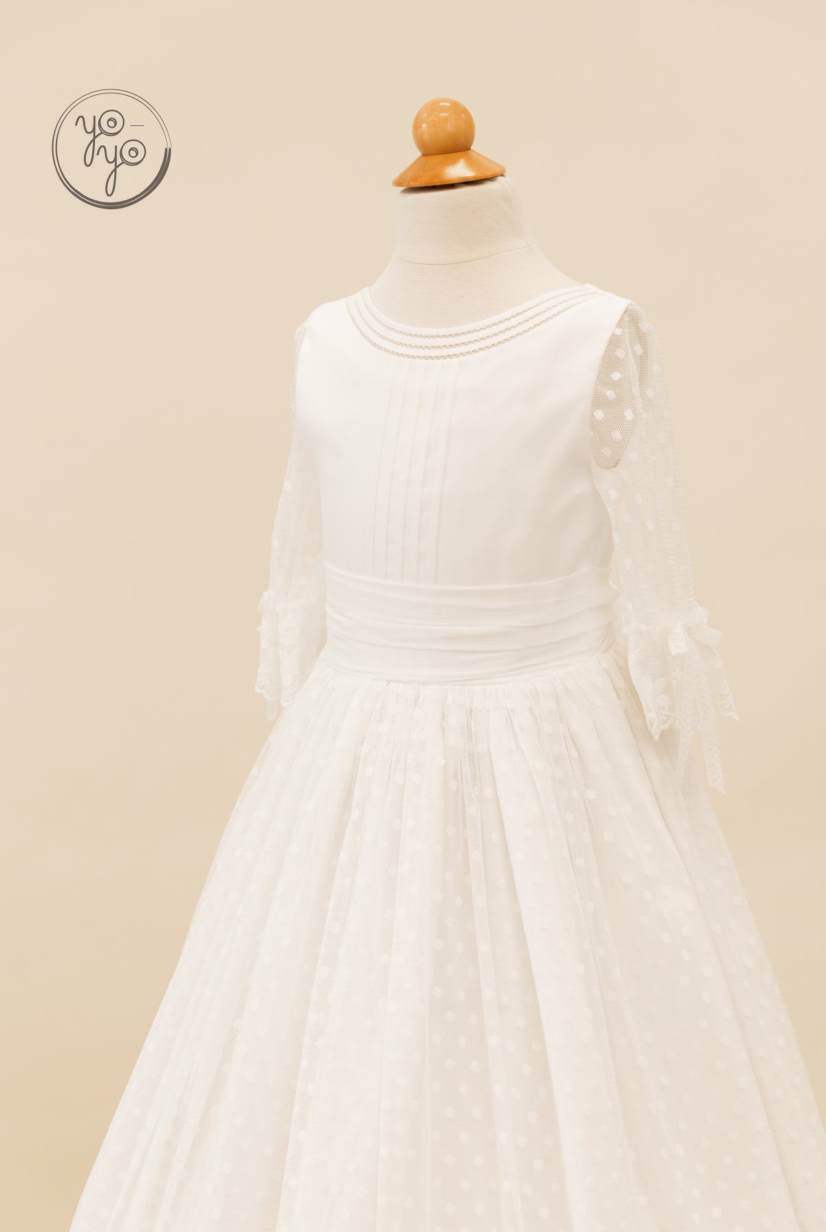 White Cassia First Communion dress featuring a pleated bodice, beaded neckline, and sheer polka-dot sleeves with tied cuffs.