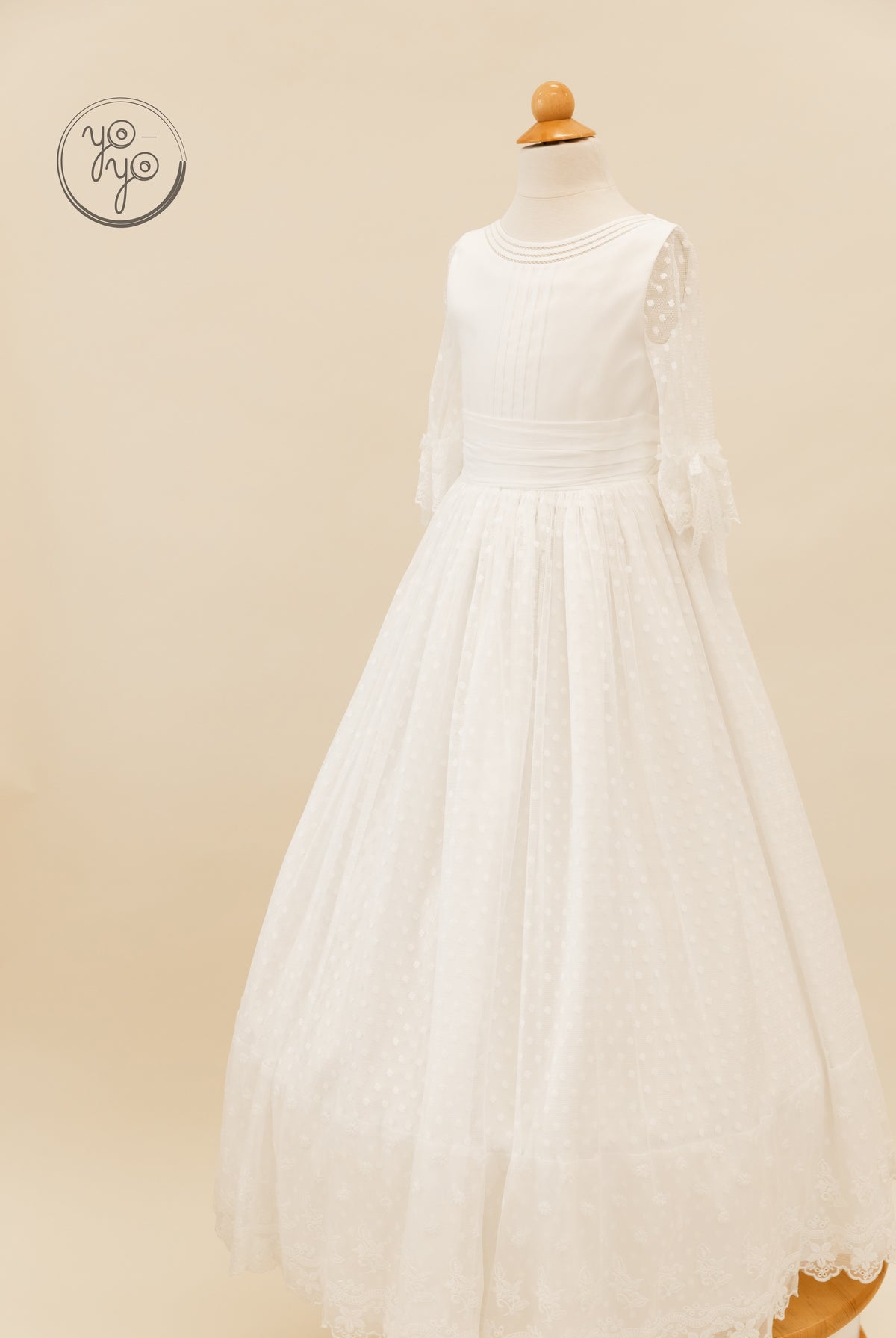YoYo Boutique Cassia white floor-length Communion dress featuring a pleated bodice, round neckline, sheer swiss-dot sleeves, and a lace hem.