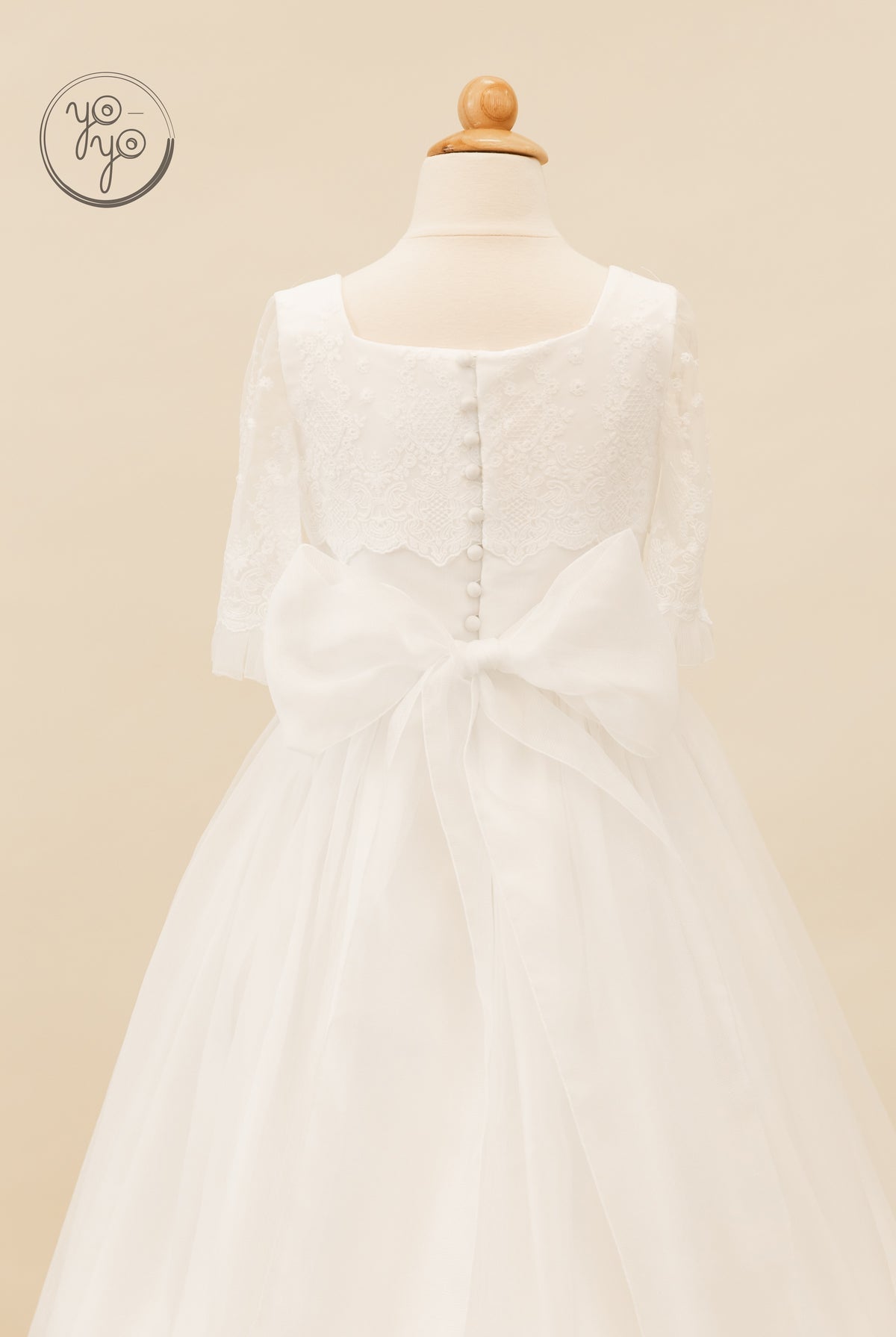 Back view of the white Campanilla dress, featuring a tea-length skirt, lace bodice, button detail, three-quarter sleeves, and an organza bow.