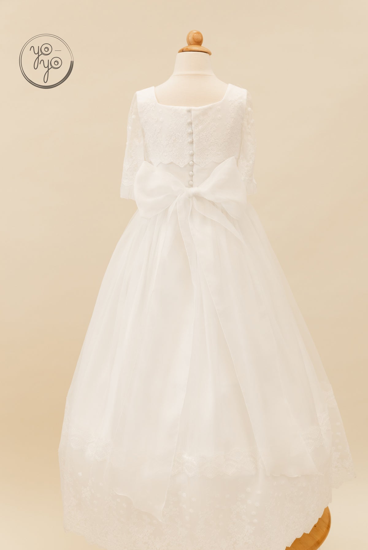 Back view of the white, tea-length Campanilla Dress by YoYo Boutique, featuring a square neckline, floral lace bodice, buttons, and an organza bow.