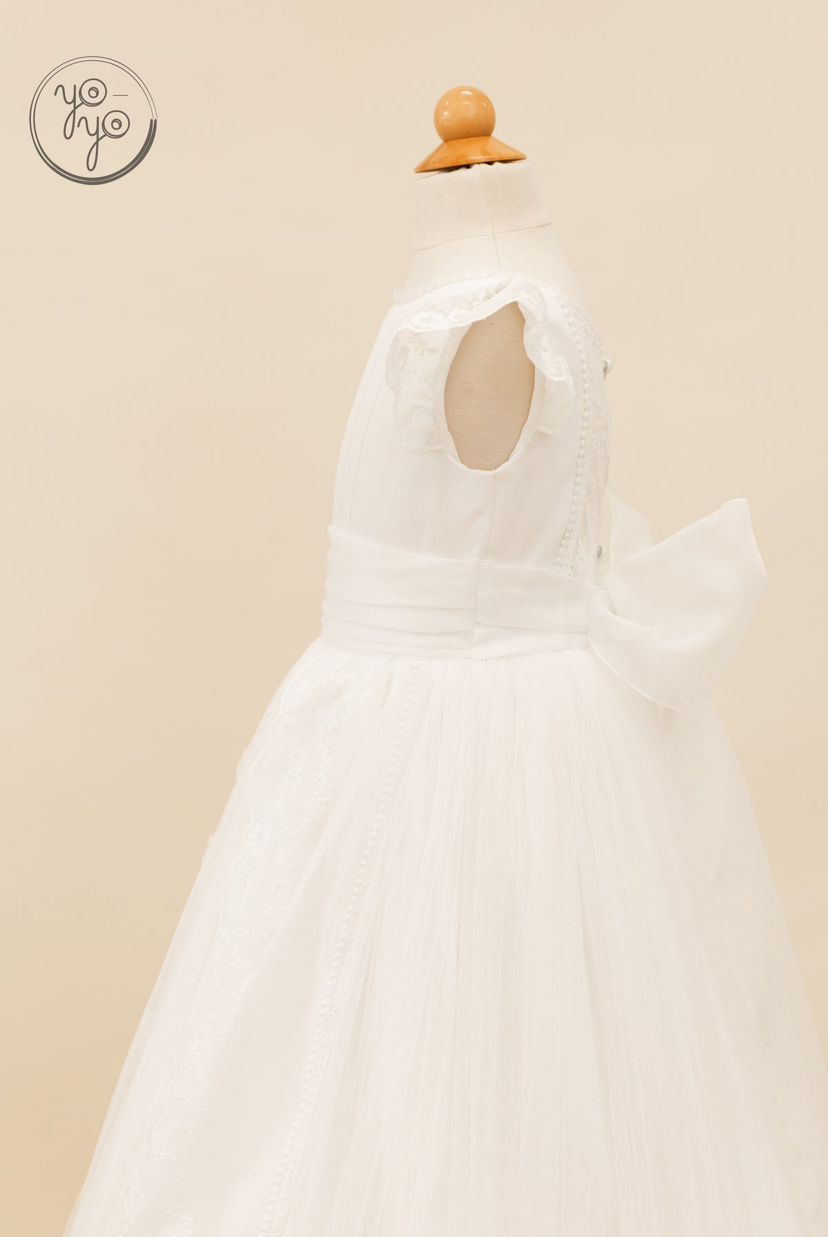 Side view of a white boho Buganvilla Dress on a mannequin, featuring lace cap sleeves, a wide waist sash, a large back bow, and a full tulle skirt.