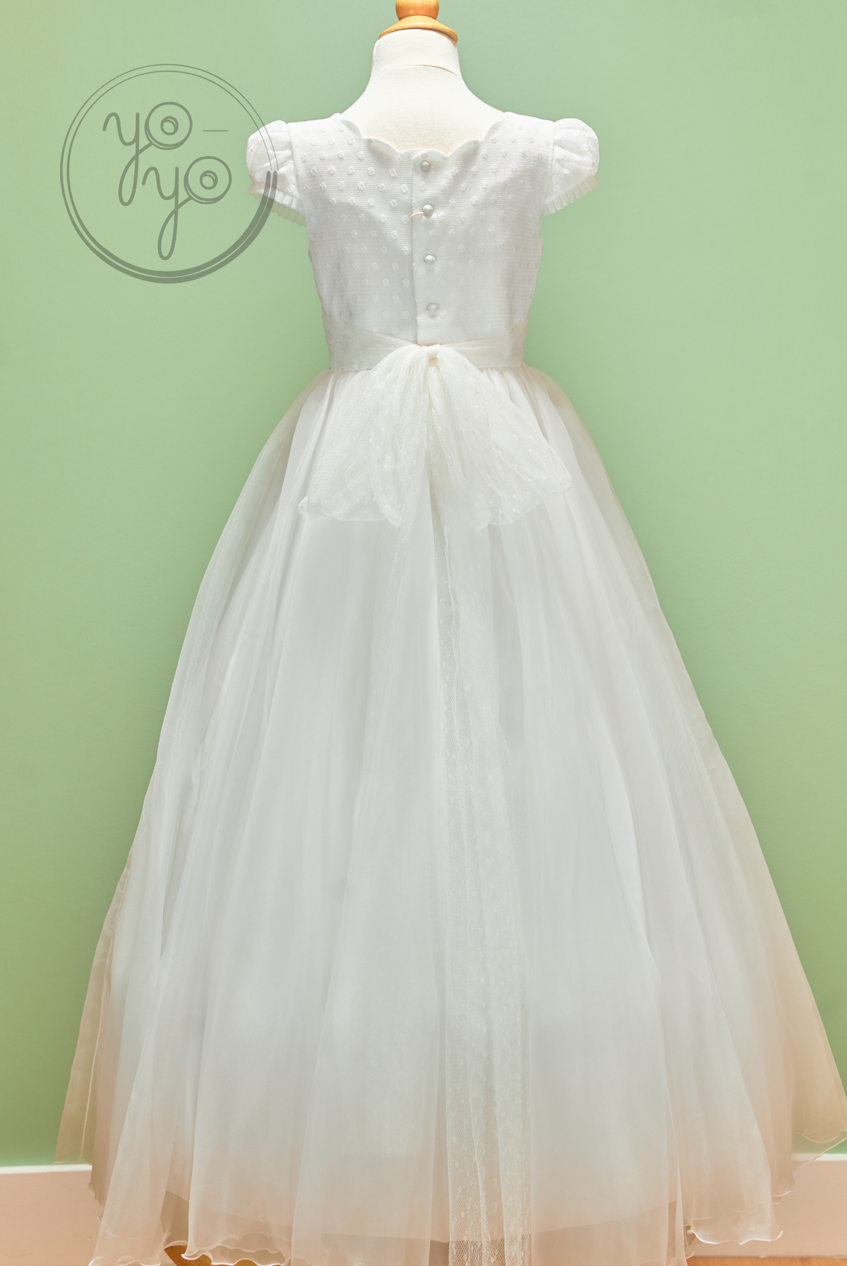 Back view of the white, floor-length Iris dress featuring puff sleeves, a button-down bodice, and a large sheer organza bow at the waist.