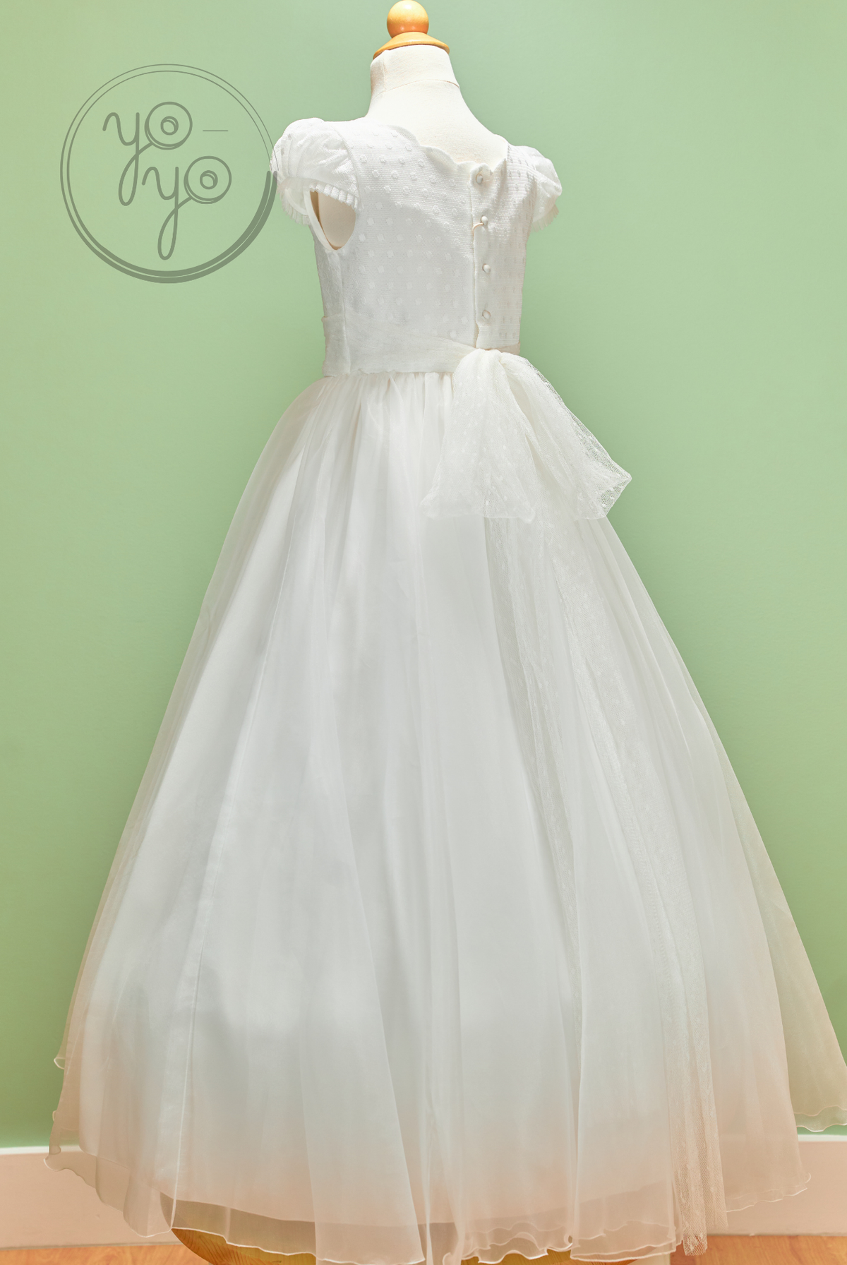 White sleeveless Iris Dress by YoYo Boutique on a mannequin, featuring a textured bodice, button-back, and embroidered tulle skirt with a large bow.
